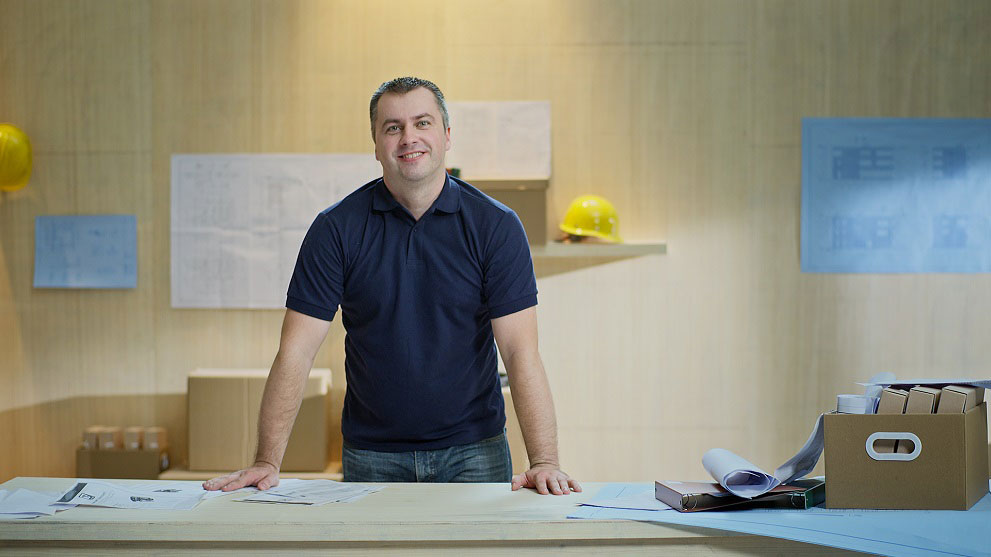 ชายยิ้มในชุดสีน้ำเงินยืนอยู่ที่โต๊ะที่มีแผนการก่อสร้างและแฟ้มเอกสาร หมวกกันน็อคสีเหลืองและแผนอยู่ในพื้นหลัง