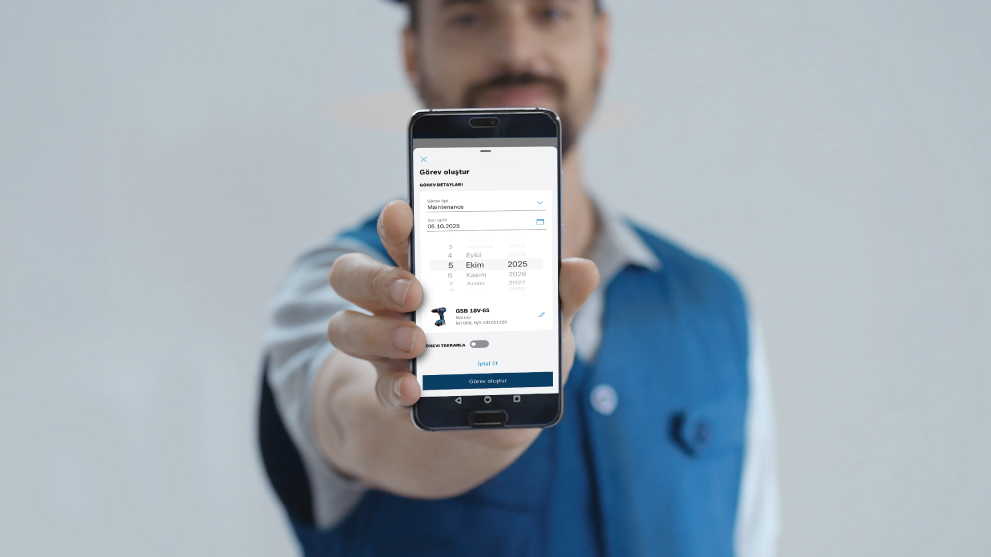 ชายคนหนึ่งแสดงสมาร์ทโฟนที่มีแอปพลิเคชันที่แสดงรายละเอียดงาน เขาสวมเสื้อเชิ้ตสีน้ำเงิน