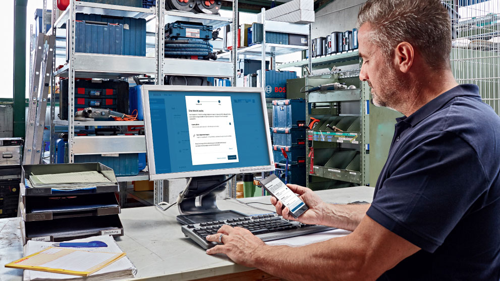 ชายคนหนึ่งกำลังทำงานที่คอมพิวเตอร์ในขณะที่ถือสมาร์ทโฟนไว้ในมือ ด้านหลังมีชั้นวางที่เต็มไปด้วยกล่องเครื่องมือและวัสดุ