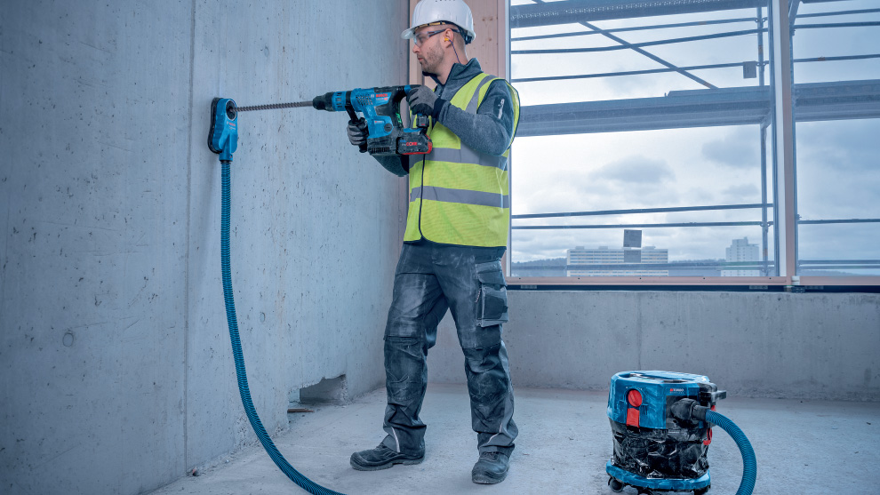 ชายคนหนึ่งสวมหมวกกันน็อคและเสื้อกั๊กกำลังเจาะผนังคอนกรีต.