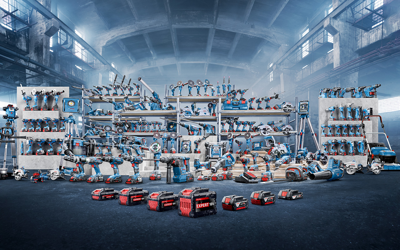 เครื่องมือ Bosch Professional สีน้ำเงินจำนวนมากถูกจัดเรียงบนชั้นวางและพื้นในโรงงานอุตสาหกรรม มีเครื่องมือไร้สาย เครื่องจักร และอุปกรณ์เสริมหลายรายการ รวมถึงสว่าน เครื่องขัด และแบตเตอรี่ที่มีคำว่า 'EXPERT' พื้นหลังมีหมอกและให้บรรยากาศอุตสาหกรรม